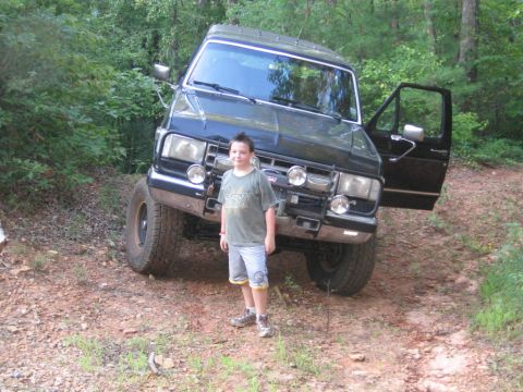 1988 Ford Bronco - A young boy stands in front of a lifted black Ford Bronco with its driver's side door open. The vehicle is parked on a dirt path surrounded…