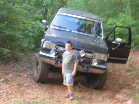 1988 Ford Bronco - A young boy stands in front of a lifted, dark-colored Ford Bronco with its driver's side door open. The vehicle is parked on a dirt path su…