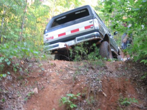 1988 Ford Bronco - The rear of a dark colored SUV is angled on a dirt path in a wooded area. A person is visible in the background to the right.