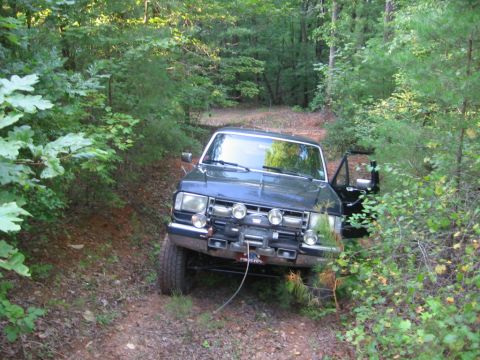 1988 Ford Bronco - A dark colored Ford Bronco is parked on a dirt trail surrounded by dense green foliage and trees. The front of the vehicle is visible, feat…