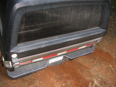1988 Ford Bronco - The rear bumper and tailgate area of a dark-colored vehicle, possibly a truck or SUV, is shown partially stuck in reddish-brown dirt. A lic…