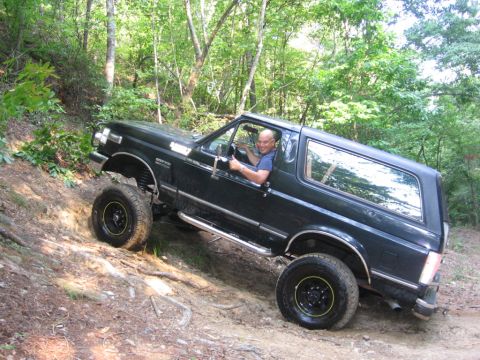 1988 Ford Bronco - A dark blue Ford Bronco is driving up a steep, dirt trail in a wooded area. The driver is visible through the open driver's side window, sm…