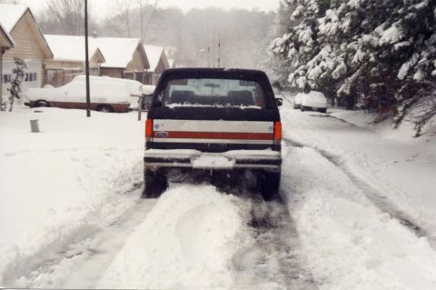 1988 Ford Bronco - The rear of a black Ford Bronco is visible, driving through deep snow on a street lined with houses and snow-covered trees.