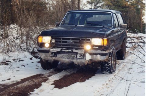 1988 Ford Bronco - A dark-colored Ford Bronco is driving through a snowy, wooded area, with its headlights on and mud and snow splashed on the front bumper an…