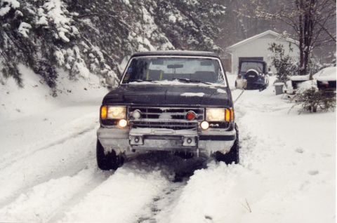 1988 Ford Bronco - A dark-colored SUV is driving through deep snow on a road lined with snow-covered trees. Another blue Jeep is visible parked in the backgro…