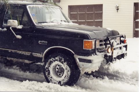 1988 Ford Bronco - A black Ford Bronco is parked in deep snow with snow falling. The front of the vehicle is covered in snow and ice, and a brush guard is vis…