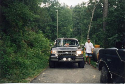 1988 Ford Bronco - A man stands next to a black Ford Bronco on a paved road surrounded by lush green trees. The front of the Bronco is visible with its headli…