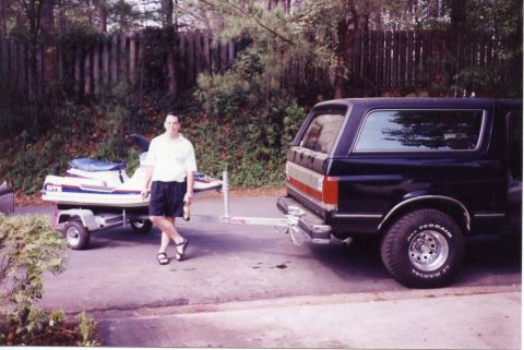 1988 Ford Bronco - A man stands next to a jet ski on a trailer, which is hitched to the rear of a dark-colored SUV. The SUV appears to be a Ford Bronco, and t…
