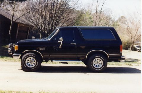 1988 Ford Bronco - The side profile of a black Ford Bronco is shown parked on a paved surface in front of a building and trees. The vehicle has chrome trim, r…