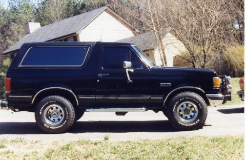 1988 Ford Bronco - A black 1988 Ford Bronco is parked on a paved surface in front of a house with a dark roof and light-colored siding. The vehicle is viewed…