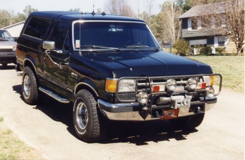 1988 Ford Bronco - A black 1988 Ford Bronco is parked on a concrete driveway in front of a house. The SUV has a brush guard with multiple lights and a winch m…