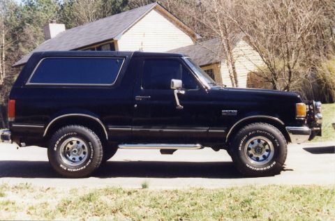 1988 Ford Bronco - A black Ford Bronco is parked on a paved surface in front of a yellow house with a dark roof. The vehicle is shown in profile with its side…