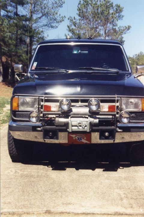 1988 Ford Bronco - Front view of a dark-colored SUV with a custom brush guard, winch, and auxiliary lights. A red and white rectangular decal with "PARAMEDIC"…