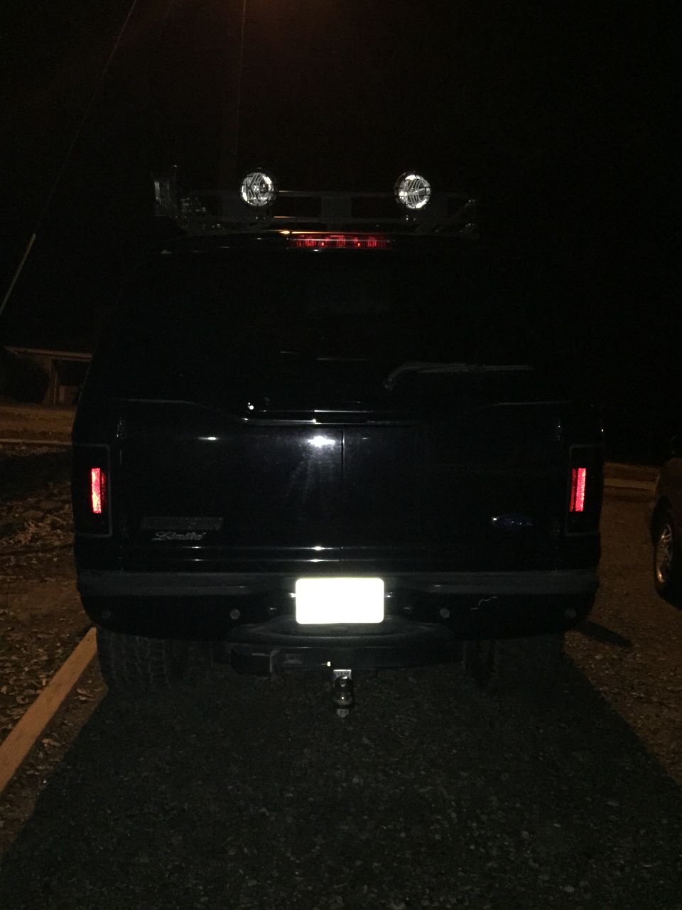 Rear view of a black SUV with roof rack spotlights and trailer hitch at night.