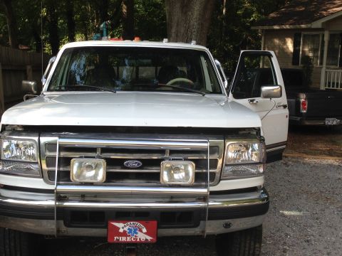 1997 Ford F-250 - The front of a white Ford F-250 pickup truck is shown with its hood up. A black pickup truck is parked behind it, and a house is visible in…
