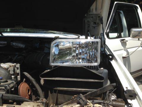 1997 Ford F-250 - A new headlight assembly is positioned over the engine bay of a white Ford F-250 pickup truck. The hood is open, revealing the engine compo…