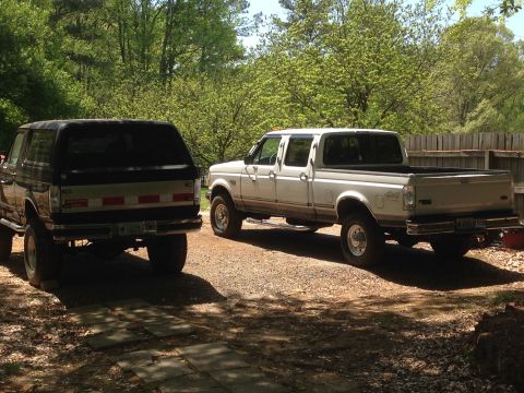 1997 Ford F-250 - Two pickup trucks are parked on a gravel driveway in front of a wooden fence and trees. The truck on the left is a dark-colored SUV with a…