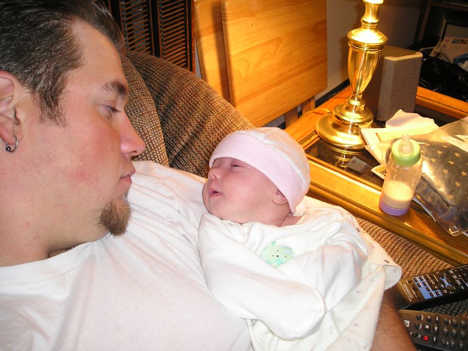 Man holding a baby on a couch, with a lamp and baby bottle on a table