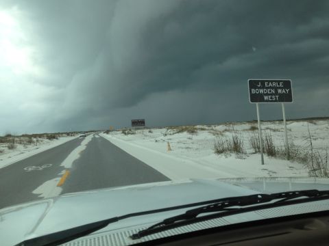 1997 Ford F-250 - The front view of a white vehicle driving on a road covered in sand or snow under a stormy sky. A sign reads "J. EARLE BOWDEN WAY WEST".