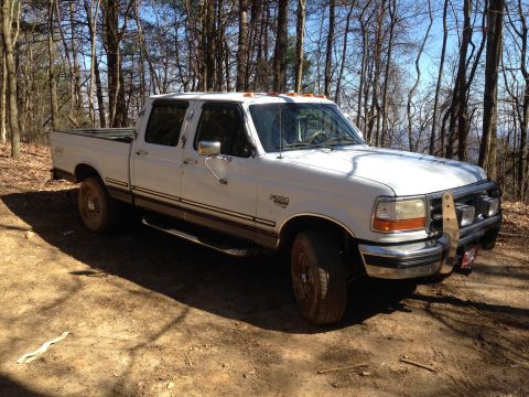 1997 Ford F-250 - A white 1990s four-door Ford F-250 pickup truck is parked on a dirt road in a wooded area. The truck has a chrome brush guard with two auxi…