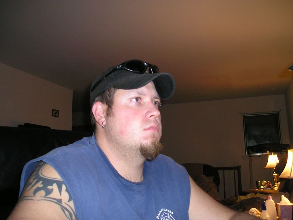 Man wearing a blue t-shirt, black baseball cap, and sunglasses, looking to the right. He has a goatee and tribal tattoos on his left arm