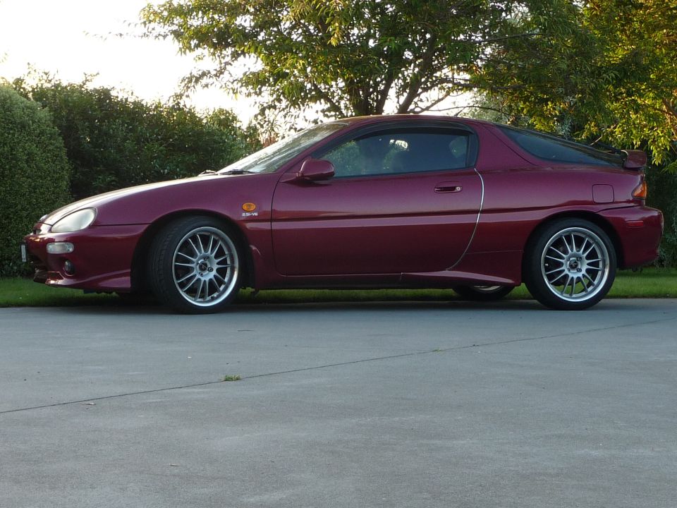 Dark red Mazda MX-3 coupe parked on a concrete driveway, side view.