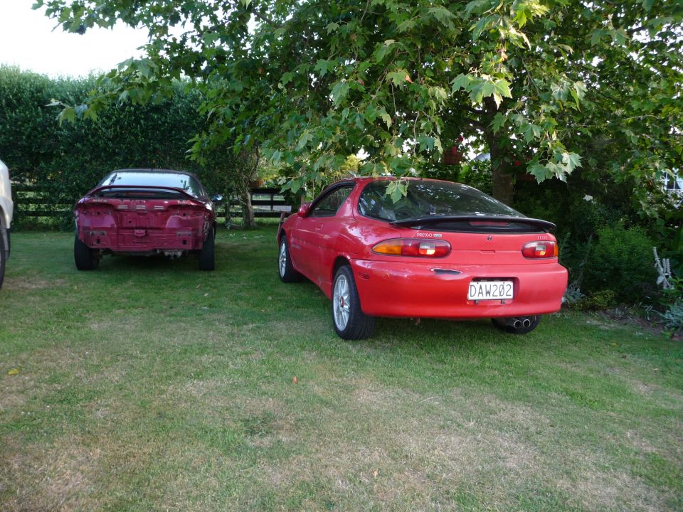 Red Mazda MX-3 parked on grass, rear view. Another red car to the left.