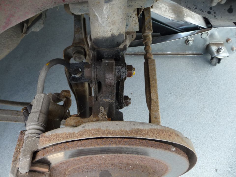 Rear suspension and brake assembly of a vehicle with a rusty brake rotor and caliper. Hydraulic jack in background.