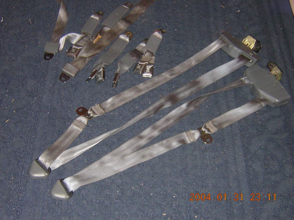 Several gray seat belts laid out on a dark surface. Some have buckles and metal clips attached