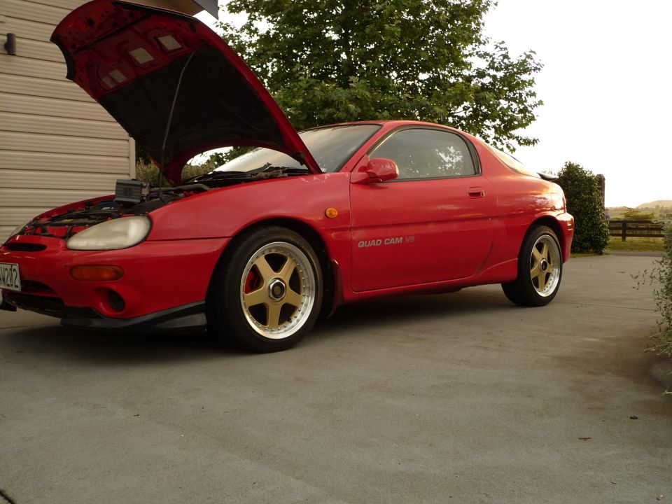 Red 1993 Mazda MX-3 coupe with hood open, gold wheels, and black spoiler.
