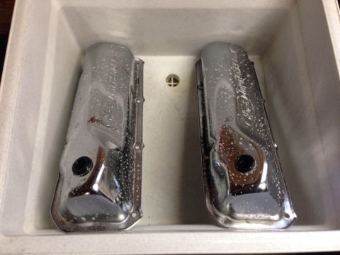 1979 Ford Bronco - Two chrome Ford valve covers are submerged in a white sink with soapy water. The valve covers have the Ford logo and text embossed on them.