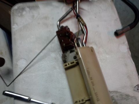 1996 Ford Bronco - A close-up view of a fuel pump assembly with a fuel level sending unit, resting on a white surface. The assembly includes a metal arm, elec…
