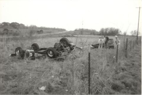 1967 Ford F-100 - A damaged truck chassis lies on its side in a grassy field, with several people gathered near another vehicle by a fence.