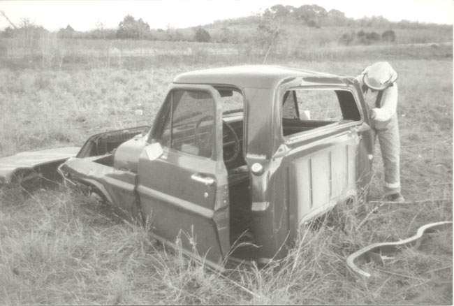 Stripped vintage Ford pickup cab shell in a field with a person working on it, black and white