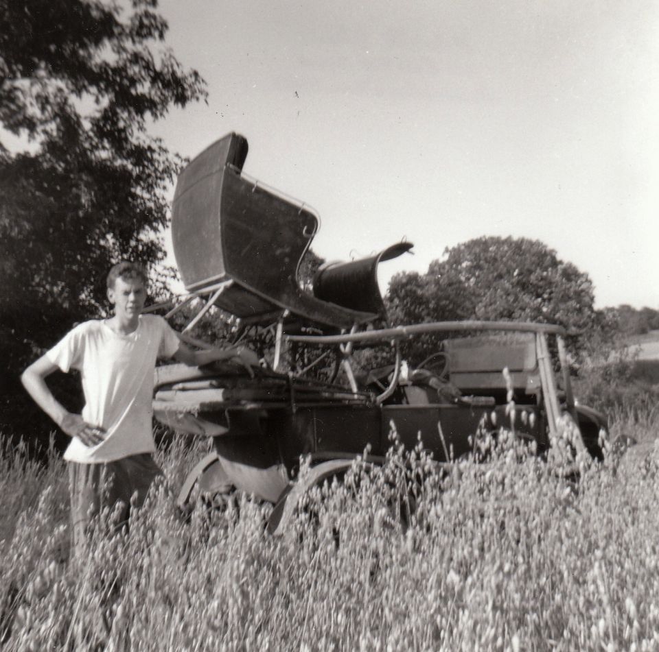 Young man in a t-shirt and shorts stands next to a vintage vehicle with a dark, carriage-like body in a grassy field.