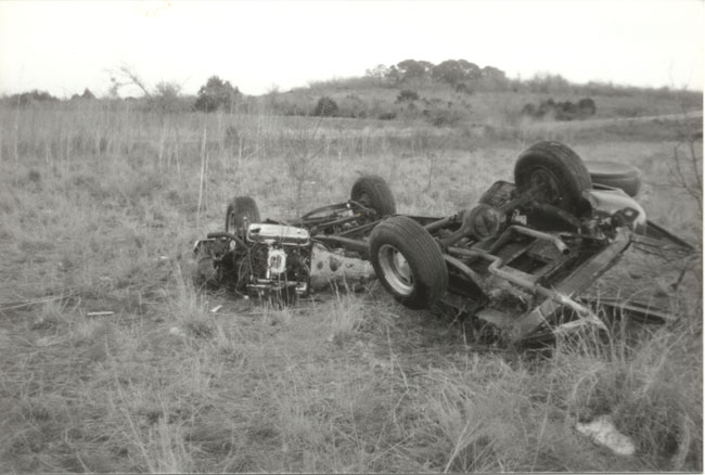 Overturned vehicle chassis in a grassy field, showing the engine and suspension