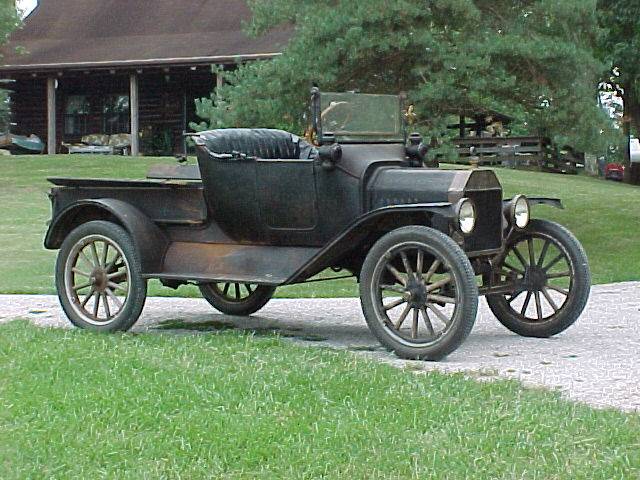 Vintage dark-colored truck with spoked wheels parked in front of a log cabin.