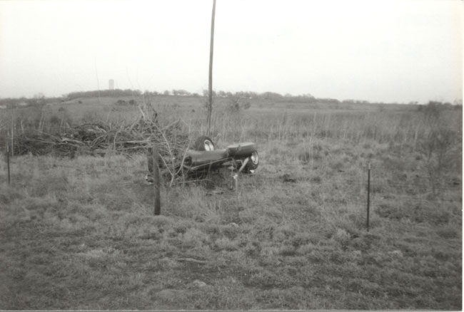 Black-and-white photo of an abandoned stripped vehicle hulk sitting in an overgrown field