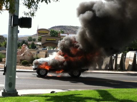 1990 Ford Bronco - A vehicle is on fire, emitting a large plume of black smoke, on a street with a "Rainbow" store in the background.