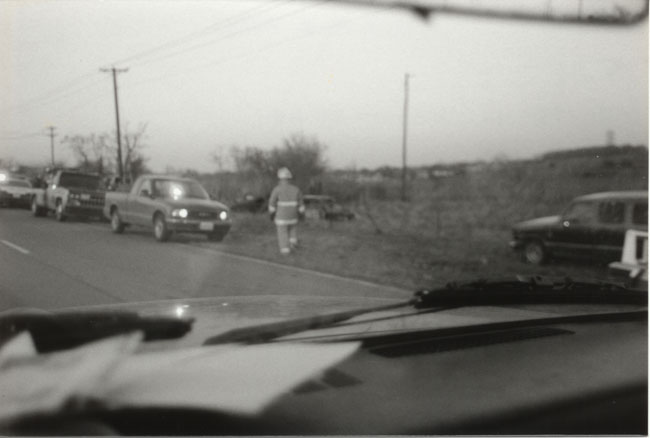 B&W photo from inside a vehicle of a firefighter walking past pickups parked along a rural roadside