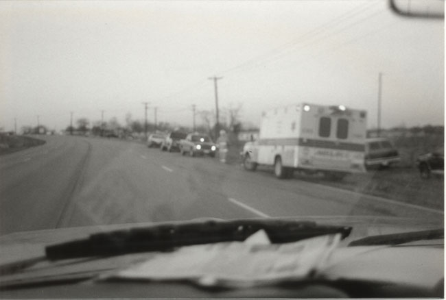 Blurry black-and-white view through a windshield of an ambulance and vehicles stopped along a rural roadside.