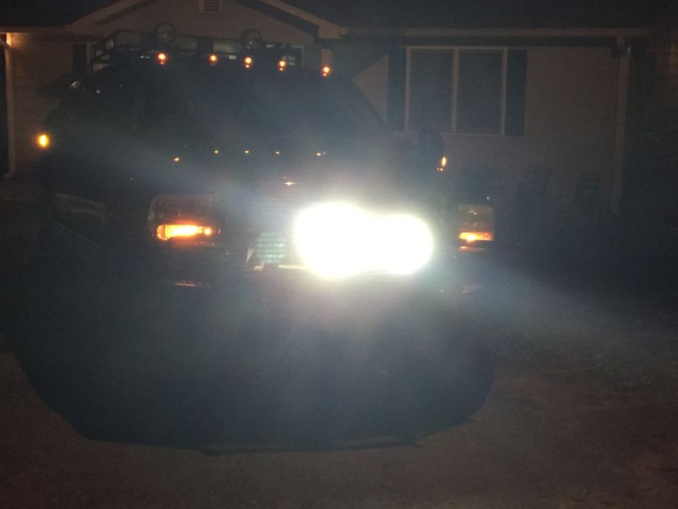 Dark-colored Ford Excursion front view at night, headlights on, roof rack with lights, in front of a house