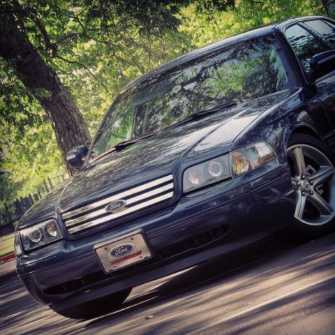 2004 Ford Crown Victoria - A dark blue Ford Crown Victoria is parked under a tree, casting shadows on the asphalt.