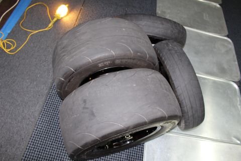2003 Ford Crown Victoria - pile of wide drag slick and treaded tires stacked on a mat and metal panels, with a yellow cord and shop light nearby.