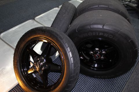 2003 Ford Crown Victoria - Two black five-spoke wheels mounted with Mickey Thompson tires arranged on a checkered mat and metal panels indoors.