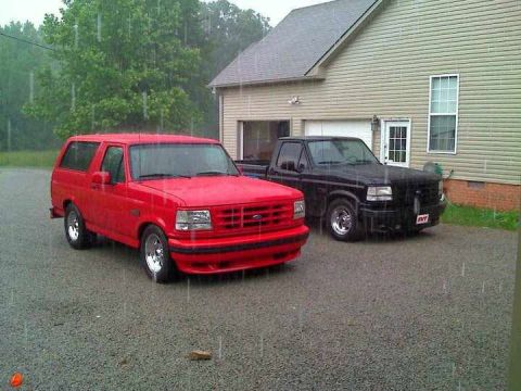 1996 Ford Bronco - Two trucks, a red Bronco and a black pickup, are parked outside a house in the rain.