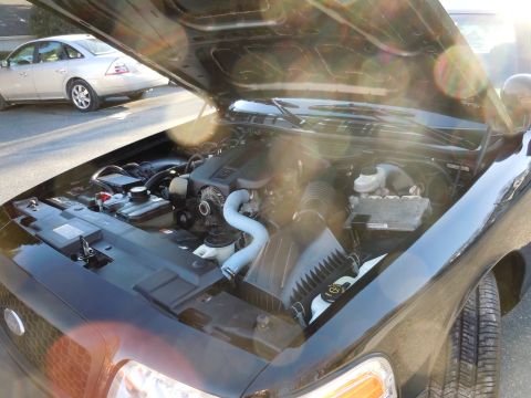 2001 Ford Crown Victoria - The engine bay of a black vehicle is shown with the hood open, while a silver sedan is visible in the background.