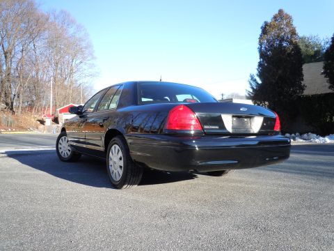 2001 Ford Crown Victoria - The rear passenger side of a black 2001 Ford Crown Victoria is shown parked on an asphalt road with trees and a building in the background.
