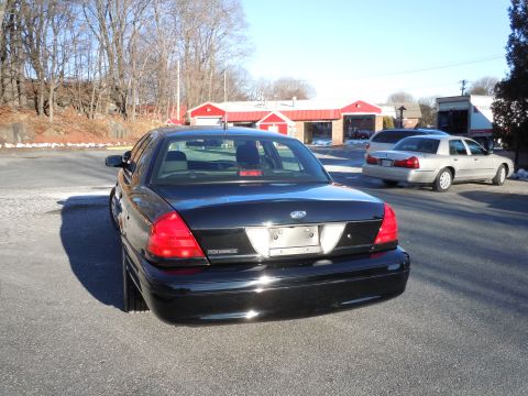 2001 Ford Crown Victoria - The rear driver's side of a black Ford Crown Victoria sedan is shown parked on asphalt. Another silver sedan is parked behind it, and a whi…