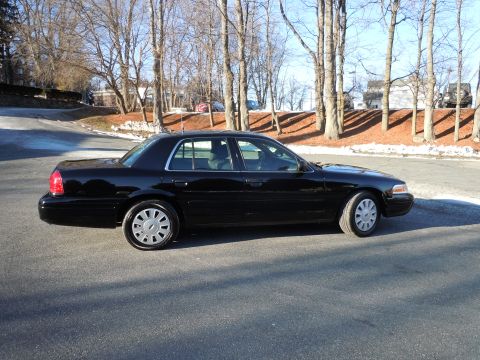 2001 Ford Crown Victoria - The side view of a black 2001 Ford Crown Victoria sedan is parked on an asphalt road with trees and buildings in the background.